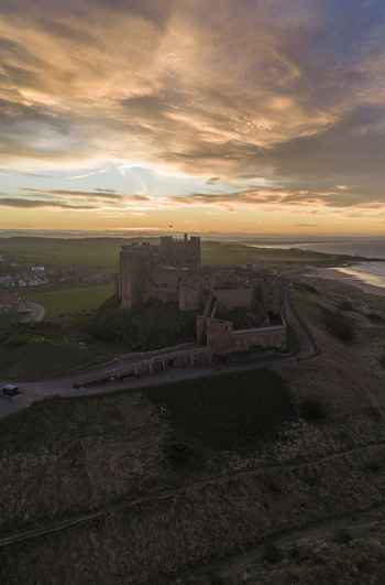 Aerial view of Bamburgh Castle standing on a coastal hill under a golden sunset sky with historic stone walls overlooking the North Sea in Bamburgh England United Kingdom. Aerial view of Bamburgh Castle standing on a coastal hill under a golden sunset sky with historic stone walls overlooking the North Sea in Bamburgh England United Kingdom.