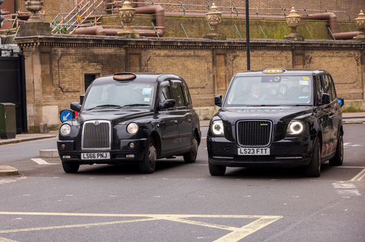 London United Kingdom 12 May 2025 View of two iconic black taxis driving along a city street past historic stone walls and decorative urns.