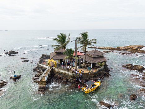 Aerial view of Seenigama Muhudu Dewalaya temple on a rocky island with palm trees people and a yellow boat in the turquoise ocean Peraliya Sri Lanka. Aerial view of Seenigama Muhudu Dewalaya temple on a rocky island with palm trees people and a yellow boat in the turquoise ocean Peraliya Sri Lanka.
