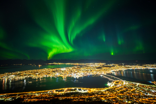 View of the city of Tromso illuminated at night under a vibrant display of northern lights in the sky above the fjord Tromso Troms Norway. View of the city of Tromso illuminated at night under a vibrant display of northern lights in the sky above the fjord Tromso Troms Norway.