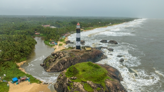 Aerial view of Kapu Light House standing on a rocky outcrop surrounded by the Arabian Sea with lush palm trees sandy coastline and crashing waves in Udupi Karnataka India.