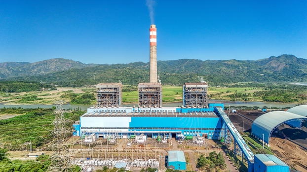 Aerial view of a large power plant with a tall chimney emitting smoke blue industrial buildings and surrounding green hills in Indonesia.