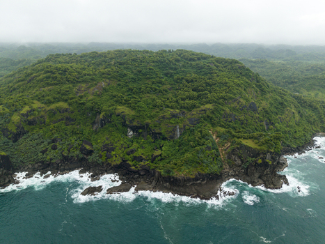Aerial view of the rugged coastline where turquoise waves crash against dark rocks beneath a verdant hill Gunungkidul Regency Daerah Istimewa Yogyakarta Indonesia. Aerial view of the rugged coastline where turquoise waves crash against dark rocks beneath a verdant hill Gunungkidul Regency Daerah Istimewa Yogyakarta Indonesia.