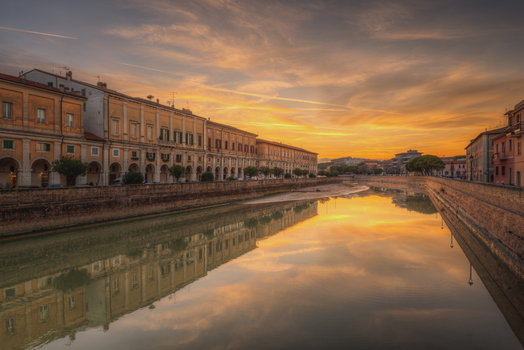 View of the Misa river reflecting the warm golden hour sky and the historic porticoed buildings along the embankment in Senigallia Marche Italy.