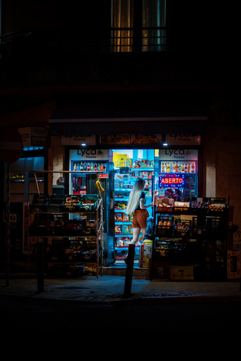 Alges Lisbon Portugal 25 July 2026 View of a woman entering a brightly lit corner store at night with produce crates displayed outside and an open sign glowing in the window.