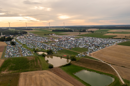 Aerial view of the Open Beatz Festival campsite featuring thousands of tents small ponds and green fields with wind turbines under a sunset sky in Herzogenaurach Bayern Germany. Aerial view of the Open Beatz Festival campsite featuring thousands of tents small ponds and green fields with wind turbines under a sunset sky in Herzogenaurach Bayern Germany.