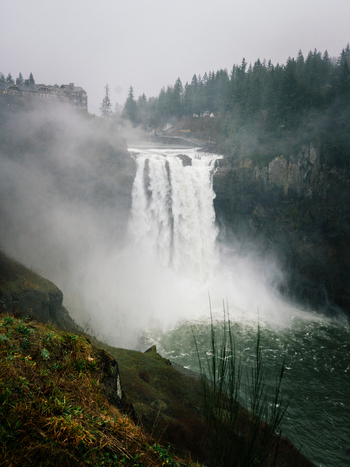 View of Snoqualmie Falls cascading over a rocky cliff with mist rising from the base surrounded by a dense evergreen forest and the Salish Lodge United States of America. View of Snoqualmie Falls cascading over a rocky cliff with mist rising from the base surrounded by a dense evergreen forest and the Salish Lodge United States of America.