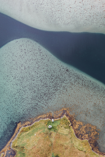 Aerial view of the coastal landscape of Lofoten featuring a small grassy peninsula with a few buildings surrounded by shallow turquoise waters in Leknes Nordland Norway. Aerial view of the coastal landscape of Lofoten featuring a small grassy peninsula with a few buildings surrounded by shallow turquoise waters in Leknes Nordland Norway.