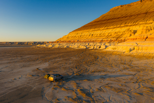 Aerial view of an off road vehicle and a tent parked near a massive layered plateau in the desert during golden hour in Mangystau Region Kazakhstan. Aerial view of an off road vehicle and a tent parked near a massive layered plateau in the desert during golden hour in Mangystau Region Kazakhstan.