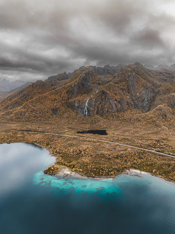 Aerial view of Lofoten landscape featuring turquoise coastal waters rugged mountains with autumn foliage a small waterfall and a winding road in Leknes Nordland Norway. Aerial view of Lofoten landscape featuring turquoise coastal waters rugged mountains with autumn foliage a small waterfall and a winding road in Leknes Nordland Norway.