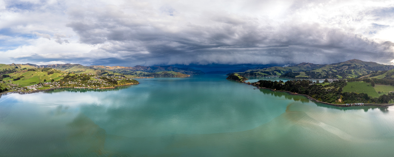 Aerial view of the bay with calm turquoise waters rolling green hills and dramatic storm clouds gathering over the landscape in Duvauchelle Canterbury Region New Zealand. Aerial view of the bay with calm turquoise waters rolling green hills and dramatic storm clouds gathering over the landscape in Duvauchelle Canterbury Region New Zealand.