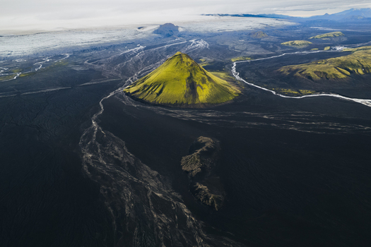 Aerial view of a vibrant green mossy symmetrical volcano rising from a stark black volcanic landscape contrasting sharply with a glacial backdrop Maelifell Iceland. Aerial view of a vibrant green mossy symmetrical volcano rising from a stark black volcanic landscape contrasting sharply with a glacial backdrop Maelifell Iceland.