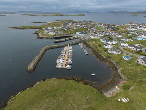 Aerial view of the harbor in Hamnavoe with moored boats at the jetty surrounded by coastal houses green hills and rocky shorelines in Hamnavoe Scotland United Kingdom. Aerial view of the harbor in Hamnavoe with moored boats at the jetty surrounded by coastal houses green hills and rocky shorelines in Hamnavoe Scotland United Kingdom.