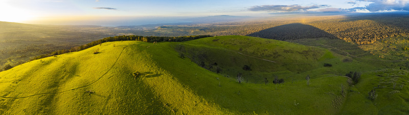 Aerial view of the lush green slopes of Puu Waawaa cinder cone at golden hour with a long shadow cast over the forest and the distant coastline Hawaii County Hawaii United States. Aerial view of the lush green slopes of Puu Waawaa cinder cone at golden hour with a long shadow cast over the forest and the distant coastline Hawaii County Hawaii United States.