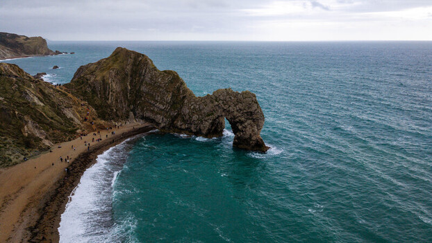Aerial view of Durdle Door a natural limestone arch on the Jurassic Coast with people on the beach and turquoise waves crashing against the shore in Wareham England United Kingdom. Aerial view of Durdle Door a natural limestone arch on the Jurassic Coast with people on the beach and turquoise waves crashing against the shore in Wareham England United Kingdom.