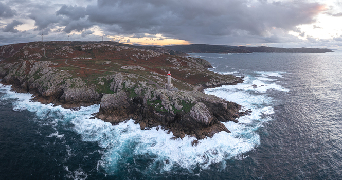 Aerial view of Faro de Punta Nariga lighthouse on a rugged rocky coastline with crashing waves and wind turbines under a cloudy sky Malpica A Coruna Spain. Aerial view of Faro de Punta Nariga lighthouse on a rugged rocky coastline with crashing waves and wind turbines under a cloudy sky Malpica A Coruna Spain.