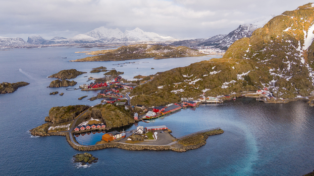 Aerial view of Lofoten featuring traditional red wooden houses nestled among rocky coastal terrain surrounded by clear blue water and snow capped mountains in Nordland Norway.