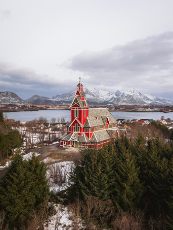 Aerial view of the vibrant red Buksnes Church standing proudly amidst a serene landscape of snow dusted trees and distant mountains Gravdal Nordland Norway. Aerial view of the vibrant red Buksnes Church standing proudly amidst a serene landscape of snow dusted trees and distant mountains Gravdal Nordland Norway.