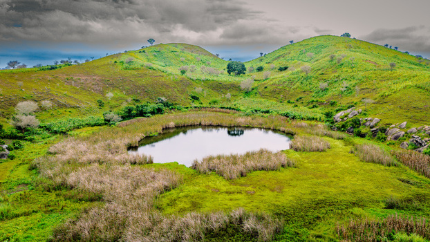 Aerial view of a serene crater lake nestled within lush green rolling hills featuring tall grasses rocky outcrops and a dramatic cloudy sky in Mangu Plateau Nigeria. Aerial view of a serene crater lake nestled within lush green rolling hills featuring tall grasses rocky outcrops and a dramatic cloudy sky in Mangu Plateau Nigeria.