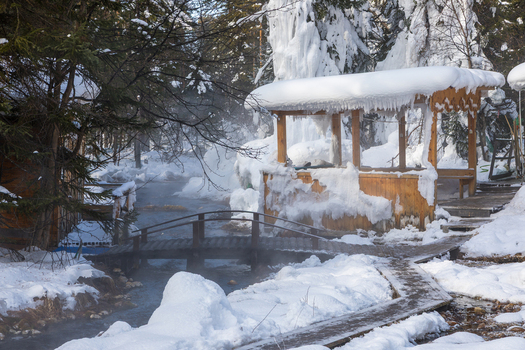 View of Goryachiye Istochniki Khakusy with a wooden bridge over a steaming thermal spring snow covered trees and a wooden gazebo in Severobaykal skiy Rayon Respublika Buryatiya Russia.