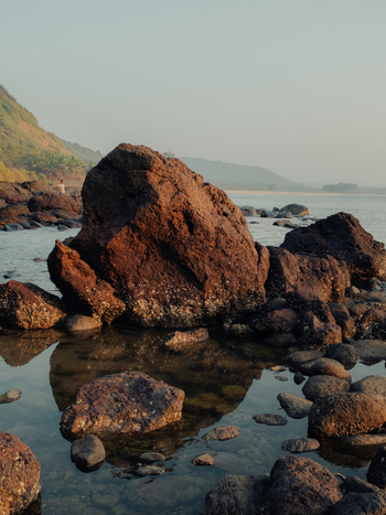 View of serene rocks reflecting in tranquil water with a distant mountain backdrop under a soft sky creating a peaceful coastal scene Chiplun Maharashtra India. View of serene rocks reflecting in tranquil water with a distant mountain backdrop under a soft sky creating a peaceful coastal scene Chiplun Maharashtra India.