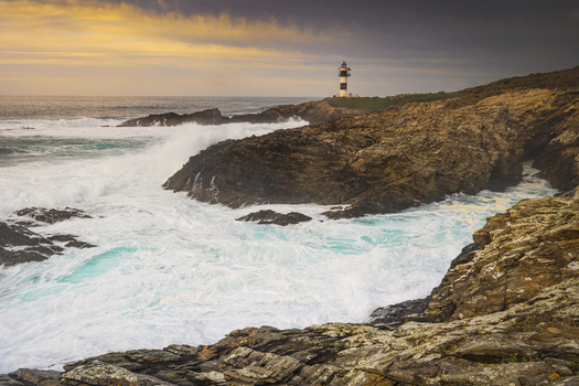 View of Illa Pancha lighthouse standing on rugged cliffs as powerful waves crash against the rocks under a golden sunset sky Lugo Lugo Spain.