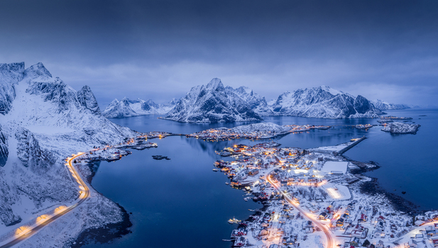 Aerial view of the snowy coastal village of Lofoten with illuminated roads and buildings nestled among dramatic mountains and deep blue waters in Nordland Norway.