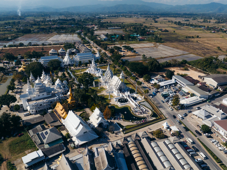Aerial view of the intricate white architecture of Wat Rong Khun temple complex with people walking on the bridge and surrounding grounds in Pa O Don Chai Chiang Rai Thailand. Aerial view of the intricate white architecture of Wat Rong Khun temple complex with people walking on the bridge and surrounding grounds in Pa O Don Chai Chiang Rai Thailand.