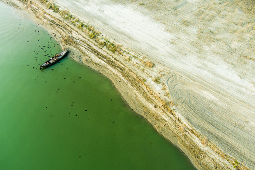 Aerial view of a small wooden boat moored near the sandy riverbank with birds flying over the green water in Rajshahi Rajshahi Division Bangladesh.