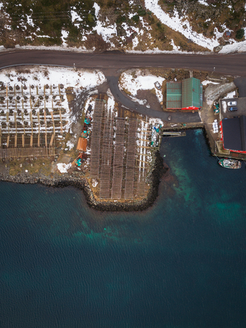 Aerial view of the rugged coastline meeting the deep blue sea with docks and buildings clustered near the harbor Ballstad Nordland Norway. Aerial view of the rugged coastline meeting the deep blue sea with docks and buildings clustered near the harbor Ballstad Nordland Norway.