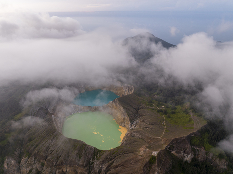 Aerial view of Kelimutu National Park with turquoise and green volcanic crater lakes surrounded by rugged mountain terrain and drifting clouds in Detusoko East Nusa Tenggara Indonesia.