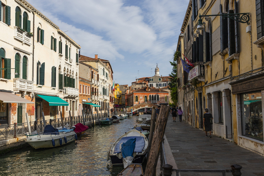 View of a narrow canal in Venice with moored boats historic buildings a stone bridge in the distance and people walking along the embankment in Venice Veneto Italy. View of a narrow canal in Venice with moored boats historic buildings a stone bridge in the distance and people walking along the embankment in Venice Veneto Italy.