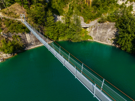 Aerial view of a metal suspension bridge spanning the turquoise waters of Lac de la Gruyere surrounded by lush green forests and rocky cliffs in Pont la Ville Fribourg Switzerland.