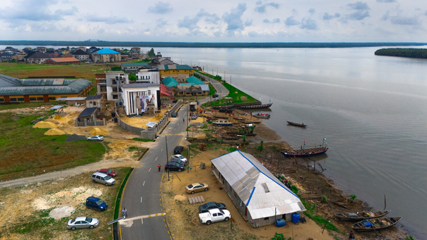 Aerial view of a coastal town with buildings a paved road parked cars and small wooden boats moored along the riverbank in Minima Rivers Nigeria.