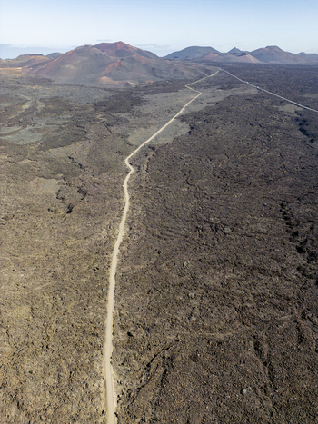 Aerial view of the volcanic landscape of Timanfaya National Park with a long dirt road cutting through the rugged lava fields in Lanzarote Canarias Spain. Aerial view of the volcanic landscape of Timanfaya National Park with a long dirt road cutting through the rugged lava fields in Lanzarote Canarias Spain.