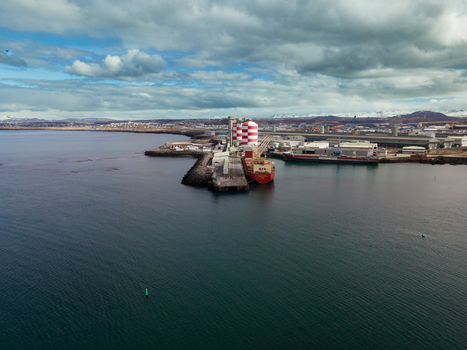 Hafnarfjordur Iceland 17 April 2026 Aerial view of the industrial port featuring a large red and white striped silo a cargo ship docked at the pier and distant snow capped mountains under a cloudy sky. Hafnarfjordur Iceland 17 April 2026 Aerial view of the industrial port featuring a large red and white striped silo a cargo ship docked at the pier and distant snow capped mountains under a cloudy sky.