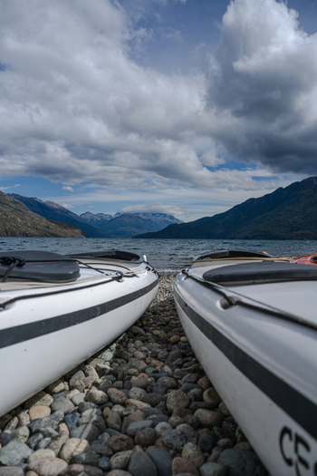 View of two white kayaks resting on a pebble beach at Lago Puelo National Park with mountains and a cloudy sky in the background in Cushamen Chubut Argentina.