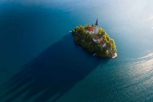 Aerial view of the Pilgrimage Church of the Assumption of Mary on Bled Island surrounded by deep blue water with long morning shadows Bled Radovljica Slovenia.