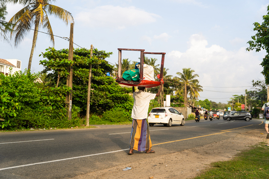 Sri Lanka 15 January 2026 View of a man carrying a wooden table on his head while walking along a roadside with palm trees and traffic.