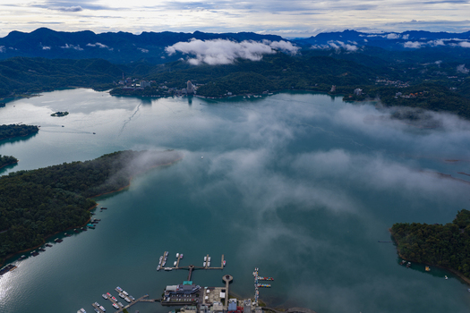 Aerial view of Sun Moon Lake with mist rolling over the turquoise water surrounding lush green mountains and boat piers in Yuchi Township Nantou County Taiwan. Aerial view of Sun Moon Lake with mist rolling over the turquoise water surrounding lush green mountains and boat piers in Yuchi Township Nantou County Taiwan.