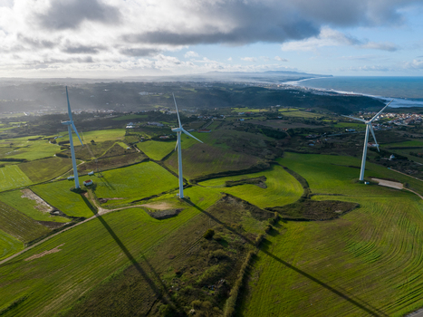 Aerial view of wind turbines standing on lush green hills overlooking the Atlantic coastline under a cloudy sky in Ribamar Portugal. Aerial view of wind turbines standing on lush green hills overlooking the Atlantic coastline under a cloudy sky in Ribamar Portugal.