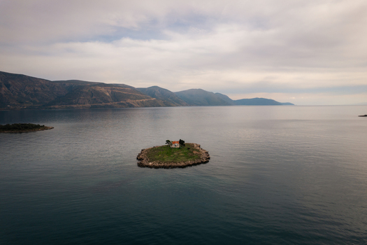 Aerial view of a small island featuring a solitary chapel surrounded by calm blue waters with distant mountains along the coastline under a soft cloudy sky in Itea Greece.