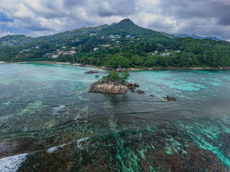 Aerial view of a small rocky island surrounded by turquoise waters contrasting with the dark seabed overlooked by lush green hills Anse Royale Seychelles. Aerial view of a small rocky island surrounded by turquoise waters contrasting with the dark seabed overlooked by lush green hills Anse Royale Seychelles.
