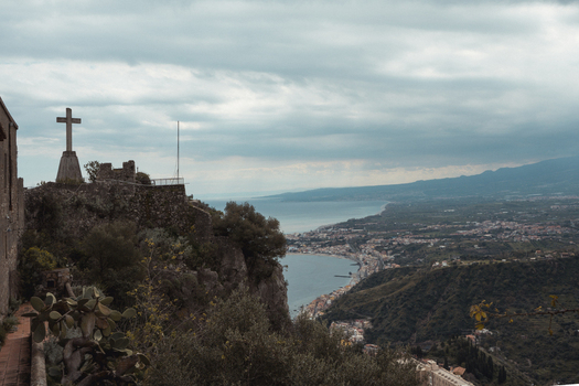 View of a stone cross perched high above the coastal town where the sea meets the land under a sky streaked with clouds Taormina Sicily Italy. View of a stone cross perched high above the coastal town where the sea meets the land under a sky streaked with clouds Taormina Sicily Italy.