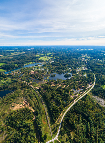 Aerial view of a lush forest landscape featuring scattered lakes wetlands and a winding road cutting through the dense greenery under a bright blue sky in Latvia. Aerial view of a lush forest landscape featuring scattered lakes wetlands and a winding road cutting through the dense greenery under a bright blue sky in Latvia.