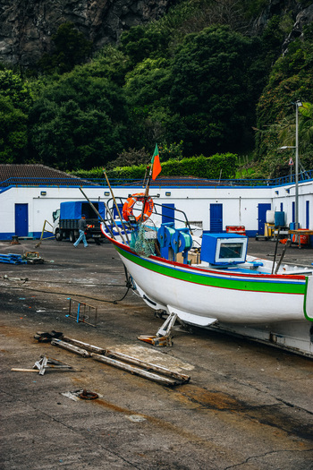 View of a vibrant fishing boat with green and red trim sits on the concrete with white buildings and lush greenery rising behind Caloura A&ccedil;ores Portugal.