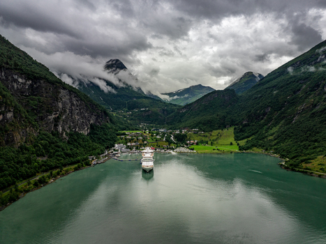 Aerial view of a large cruise ship docked in the deep waters of Geirangerfjord surrounded by steep green mountains and dramatic clouds in Geiranger More og Romsdal Norway. Aerial view of a large cruise ship docked in the deep waters of Geirangerfjord surrounded by steep green mountains and dramatic clouds in Geiranger More og Romsdal Norway.