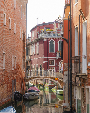View of a narrow canal lined with historic brick buildings and a small stone bridge with moored boats in the calm water in Venice Veneto Italy. View of a narrow canal lined with historic brick buildings and a small stone bridge with moored boats in the calm water in Venice Veneto Italy.