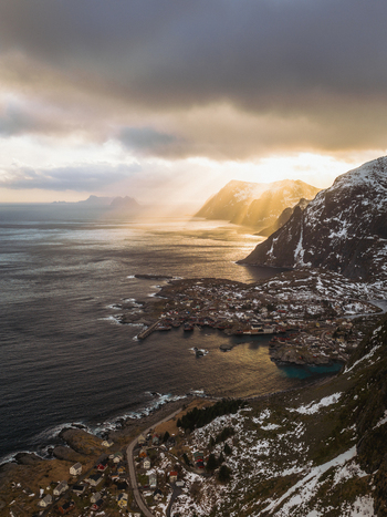 Aerial view of sun rays pierce through dramatic clouds over the snow dusted mountains and the tranquil waters of the Lofoten Islands Tind Nordland Norway.