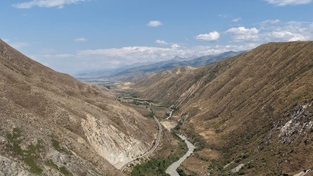 Aerial view of a winding river flowing through a deep mountain valley with rugged slopes and a road following the riverbank under a blue sky Tokmak Chuy Province Kyrgyzstan. Aerial view of a winding river flowing through a deep mountain valley with rugged slopes and a road following the riverbank under a blue sky Tokmak Chuy Province Kyrgyzstan.